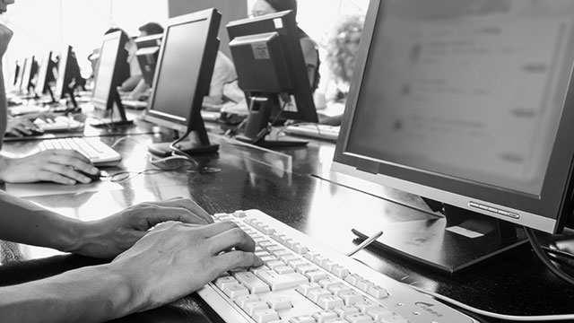 People working at desktop computers in a computer lab or office setting, with a close-up of hands typing on a keyboard in the foreground