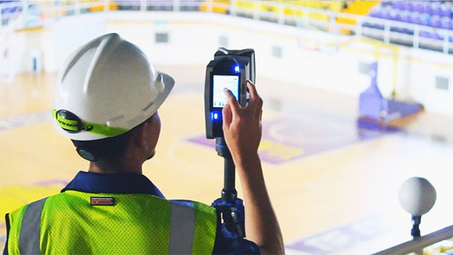 PMI Worker in safety gear using a handheld 3D laser scanner to capture spatial data inside an indoor sports facility