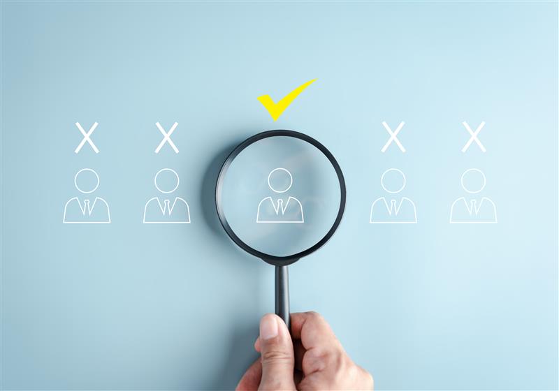 Hand holding magnifying glass highlighting a selected candidate among multiple options, with a yellow check mark indicating successful recruitment or hiring
